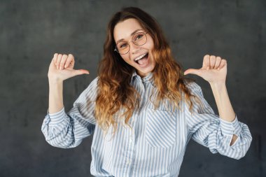 Gözlüklü, mutlu, çekici bir kadının resmi. Gri duvarın üzerinde kendini gösteren ve gülümseyen başparmaklar.