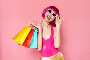 Dilini dışarı çıkaran peruk takan ve pembe arka planda izole edilmiş alışveriş torbaları tutan eğlenceli bir kız resmi.