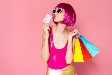 Renkli alışveriş çantaları ve pembe arka planda izole edilmiş kredi kartıyla peruk takan mutlu genç bir kız resmi.