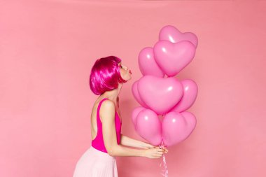 Peruklu, balonlu ve öpüşen dudakları pembe arka planda yalıtılmış genç bir kadın resmi.