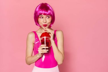 Pembe arka planda izole edilmiş plastik bardaktan soda içen peruklu güzel bir kadın görüntüsü