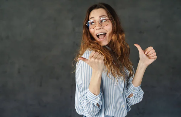 戴着眼镜的漂亮女人笑着 把大拇指伸出一边 隔着灰色的墙壁 一副快乐迷人的样子 — 图库照片