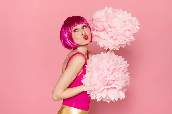 Image of amusing woman in wig making kiss lips and dancing with pom-poms isolated over pink background