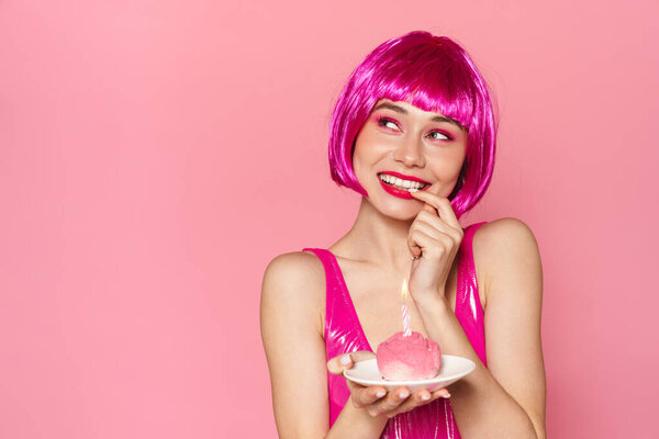 Изображение возбужденной милой женщины в парике, улыбающейся и держащей торт со свечой на розовом фоне

