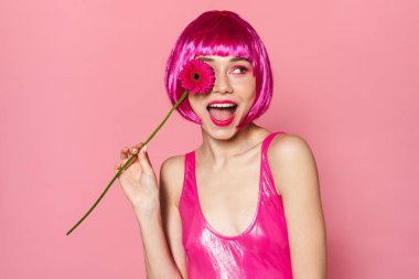 Pembe arka planda saklanmış çiçekle poz veren peruklu neşeli güzel kadın görüntüsü