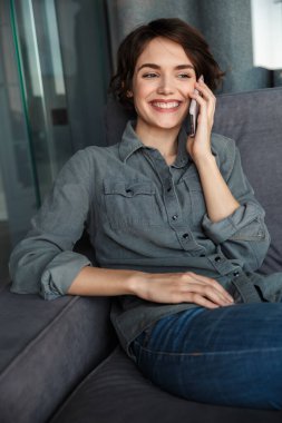 Oturma odasında kanepede otururken cep telefonuyla konuşan ve gülümseyen genç ve güzel bir kadın resmi.