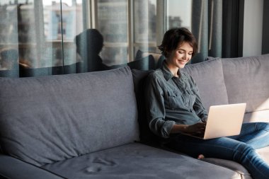 Oturma odasında kanepede otururken dizüstü bilgisayar kullanan ve gülümseyen güzel bir genç kadın görüntüsü.