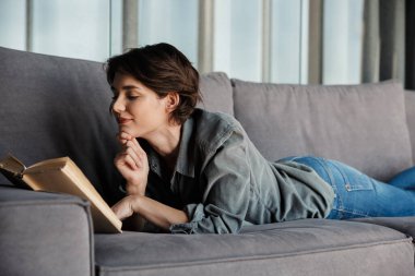 Esmer, tatlı, memnun bir kadının kitap okuyup oturma odasındaki kanepede dinlenirken gülümsemesi.