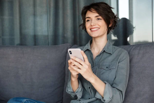 照片中美丽的年轻快乐的女人坐在客厅沙发上 用手机笑着 — 图库照片