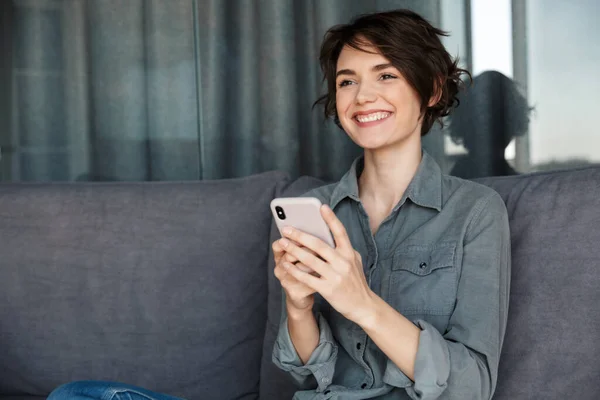 照片中美丽的年轻快乐的女人坐在客厅沙发上 用手机笑着 — 图库照片