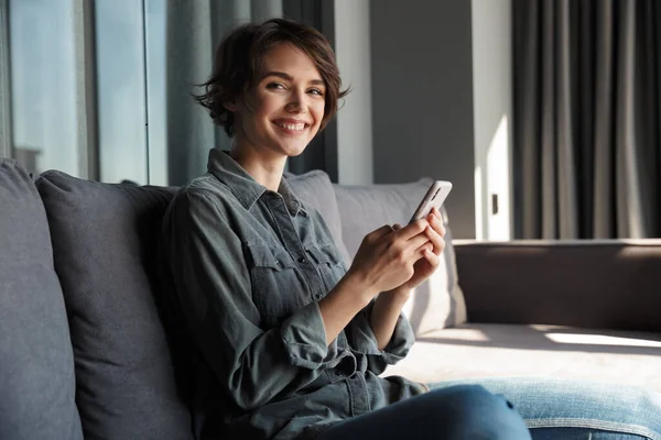 リビングルームでソファに座りながら 携帯電話と笑顔で美しい若い喜びの女性のイメージ — ストック写真