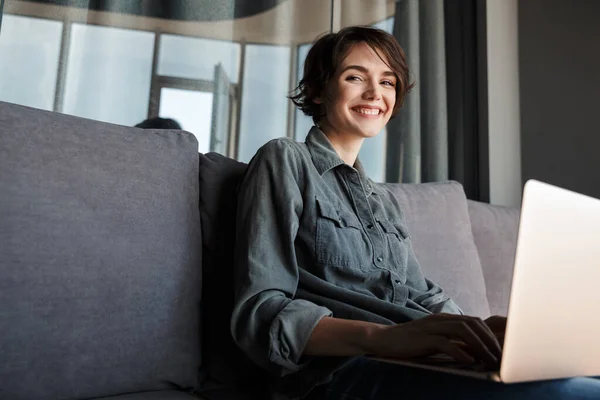 Imagem Agradável Jovem Mulher Satisfeita Usando Laptop Sorrindo Enquanto Sentado — Fotografia de Stock