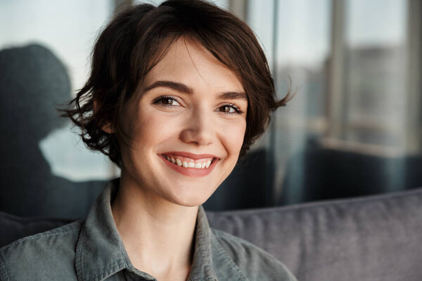 Image of brunette cute happy woman smiling and looking at camera while sitting on sofa at living room