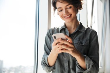 Güzel, genç ve neşeli bir kadının cep telefonu kullanması ve pencerenin yanında durup gülümsemesi.