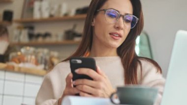 Gözlüklü çekici bir kadın beyaz bir kafede akıllı telefonunu ve dizüstü bilgisayarını kullanıyor.