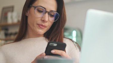 Konsantre olmuş güzel bir kadın, beyaz rahat kafede otururken akıllı telefonundan bir şey izliyor.