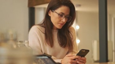 Gözlüklü hoş bir kadın akıllı telefonunu kapalı bir kafede otururken kullanıyor.