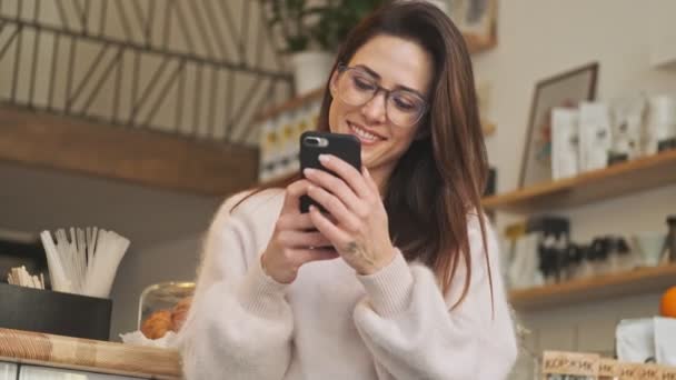 Μια Χαμογελαστή Γυναίκα Χρησιμοποιεί Smartphone Της Στο Ζεστό Καφέ Εσωτερικούς — Αρχείο Βίντεο