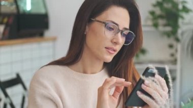 Gülümseyen, gözlüklü mutlu bir kadın kapalı bir kafede otururken akıllı telefonunu kullanıyor.