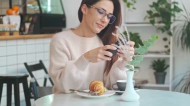 Gözlüklü güzel bir kadın kafeteryada otururken akıllı telefonuyla yemek fotoğrafı çekiyor.
