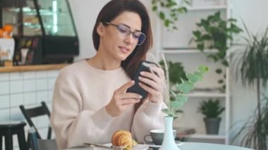 Sakin, gözlüklü bir kadın, kafeteryada otururken kruvasan yerken akıllı telefonunu kullanıyor.