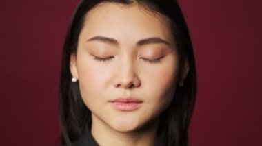Güzel, sakin, sıradan kıyafetler giyen Asyalı bir kadının yakın plan görüntüsü kırmızı arka planda tek başına gözlerini açıyor.
