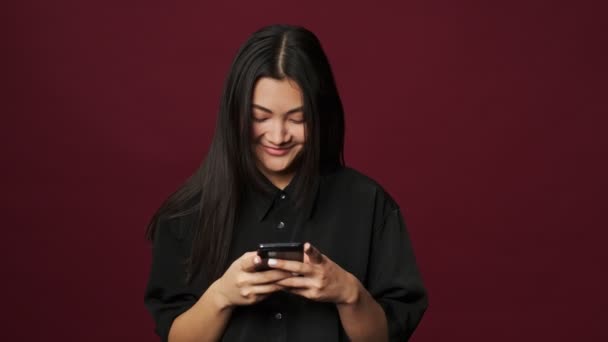 カジュアルな服を着た笑顔のアジア系の若い女性が 赤い背景に隔離されたスマートフォンを使っている — ストック動画