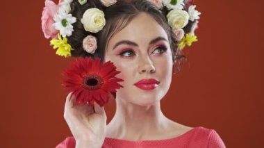 Çok hoş bir esmer kadın, inanılmaz çiçekli bir saç stiliyle elinde kırmızı bir çiçekle stüdyoda kırmızı bir arka planda poz veriyor.