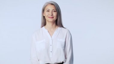 Uzun gri saçlı, resmi iş kıyafetleri giyen yaşlı ve güzel bir kadın stüdyoda beyaz arka planda izole bir şekilde gülümsüyor.