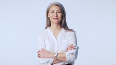 Uzun gri saçlı, mutlu ve olgun bir kadın resmi iş kıyafetleri giyerek gülüyor ve beyaz arka planda tek başına stüdyoya bakıyor.
