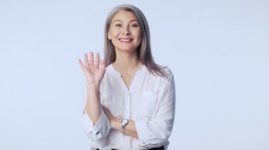 Resmi iş kıyafetleri giyen, yaşlı ve çekici bir kadın elini sallıyor. Stüdyoda beyaz arka planda tek başına duruyor.