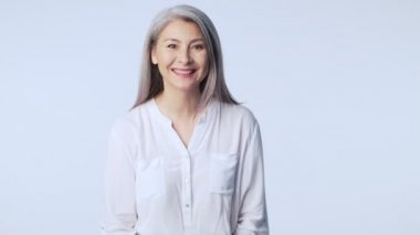 Uzun gri saçlı, mutlu bir yaşlı kadın resmi iş kıyafetleri giyerek stüdyoda beyaz arka planda tek başına dikilirken gülüyor.