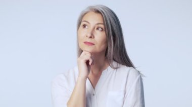 Uzun gri saçlı sakin yaşlı bir kadın resmi iş kıyafetleri giyiyor ve stüdyoda beyaz arka planda izole bir şekilde kameraya bakıyor.