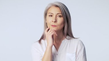 Kafası karışmış, uzun gri saçlı, resmi iş kıyafetleri giyen yaşlı bir kadın stüdyoda beyaz arka planda izole edilmiş bir şey düşünüyor.