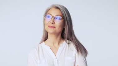 Uzun gri gözlüklü, olgun, güzel bir kadın stüdyoda beyaz arka planda tek başına duran bir şeyi düşünürken yana bakıyor.