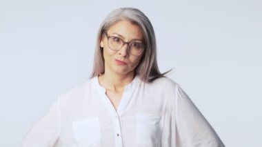 Uzun gri saçlı, kafası karışmış yaşlı ve hayal kırıklığına uğramış bir kadın stüdyoda beyaz arka planda elini kaldırıyor.
