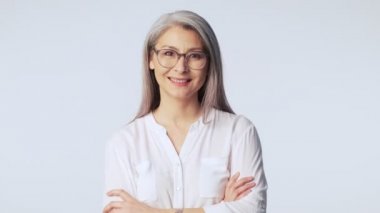 Uzun gri gözlüklü, kendinden emin, olgun bir kadın ellerini çapraz tutarak stüdyoda beyaz arka planda duran, iyi ve olumlu bir şekilde başını sallıyor.