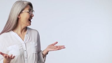Şaşırmış, kendine güvenen, uzun gri saçlı, gözlüklü yaşlı bir kadın boş bir yer göstererek stüdyoda beyaz arka planda izole edilmiş baş parmak hareketi yapıyor.