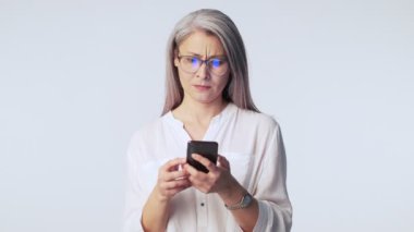 Ciddi, yaşlı, gri saçlı, gözlüklü bir kadın akıllı telefonunu kullanırken stüdyoda beyaz arka planda tek başına ayakta durmakta sorun yaşıyor.