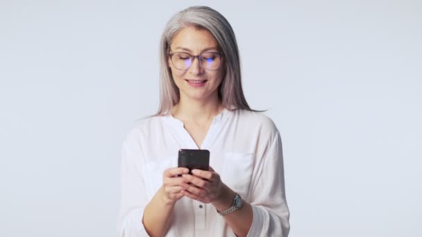 長い白髪の眼鏡をかけた笑顔の老女が スタジオで白い背景に隔離されたスマートフォンを使っている — ストック動画