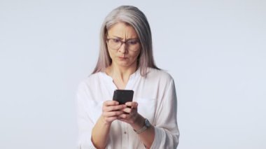 Uzun gri gözlüklü kızgın yaşlı bir kadın akıllı telefonuna yazı yazıyor ya da stüdyoda beyaz bir arkaplanda izole edilmiş birine mesaj yazıyor.