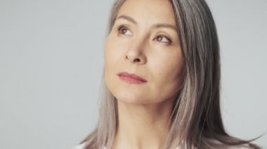 Uzun gri saçlı, rüya gören yaşlı bir kadının yakın plan görüntüsü stüdyoda beyaz arka planda izole edilmiş kameraya bakıyor.