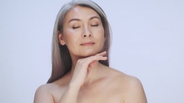 Muhteşem yaşlı, olgun, yarı çıplak bir kadın, uzun gri saçlı, poz veriyor ve mükemmel tenine dokunuyor. Stüdyodaki beyaz arka planda izole edilmiş.