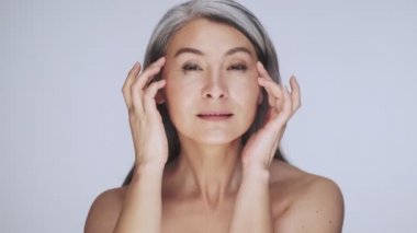 Memnun, yaşlı, yarı çıplak, uzun gri saçlı bir kadın beyaz arka planda yüzüne masaj yapıyor.