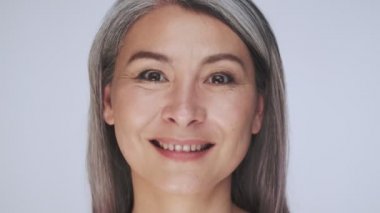 Mutlu, yaşlı, yarı çıplak, uzun gri saçlı bir kadının yakın plan görüntüsü kameraya bakıyor ve beyaz arka planda izole bir şekilde gülümsüyor.