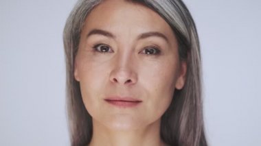 Sakin, yaşlı, yarı çıplak, uzun gri saçlı bir kadının yakın plan görüntüsü stüdyodaki beyaz arka planda izole edilmiş kameraya bakıyor.