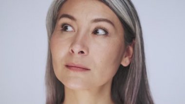 Sakin, yaşlı, yarı çıplak, uzun gri saçlı bir kadının yakın plan görüntüsü stüdyoda beyaz arka planda izole edilmiş kameraya bakıyor.