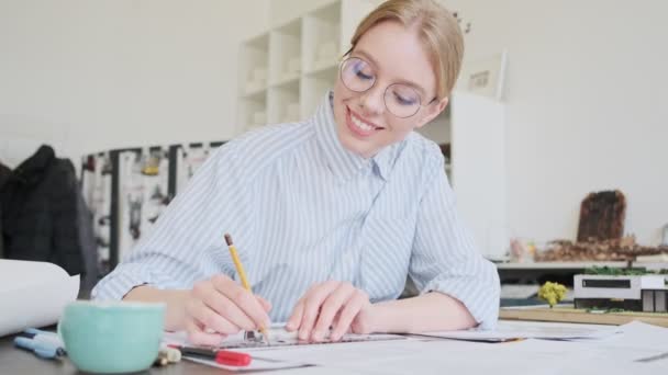 ガラスを身に着けている幸せな若い女性建築家が職場で定規を使って働いています — ストック動画
