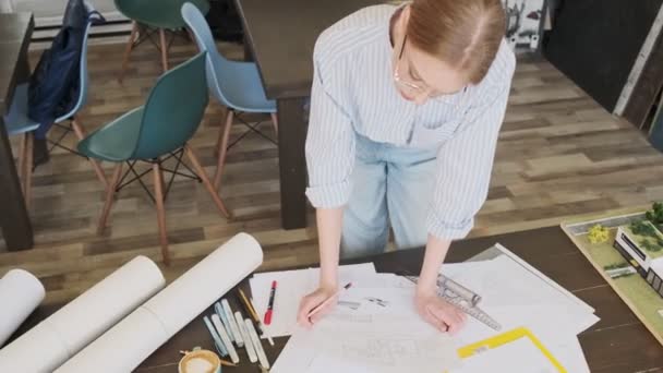 オフィスに座っている間に 若いブロンドの女性建築家が眼鏡をかけているのトップビューは 青写真の上に床計画と協力しています — ストック動画