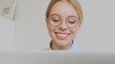 Gülümseyen, sarışın, gözlüklü genç bir mimarın yakından görüntüsü modern ofiste dizüstü bilgisayarıyla çalışıyor.